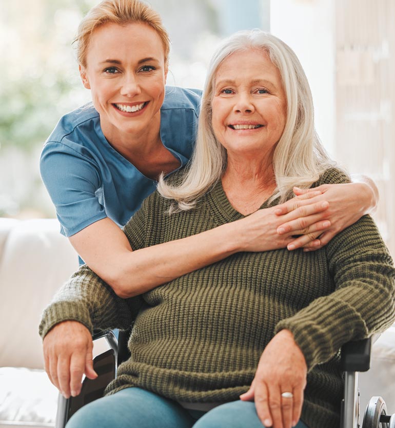 senior-healthcare Smiling caregiver in blue scrubs embracing a senior woman seated in a wheelchair indoors.