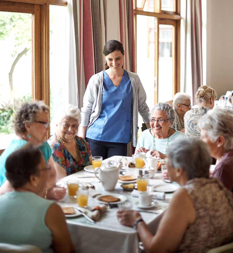 senior-assisted-living-dining A caregiver in blue scrubs and seniors enjoy breakfast together at a well-lit senior living facility.