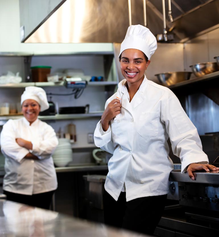 senior-living-chef Two female chefs smiling and standing in a professional kitchen with stainless steel appliances.
