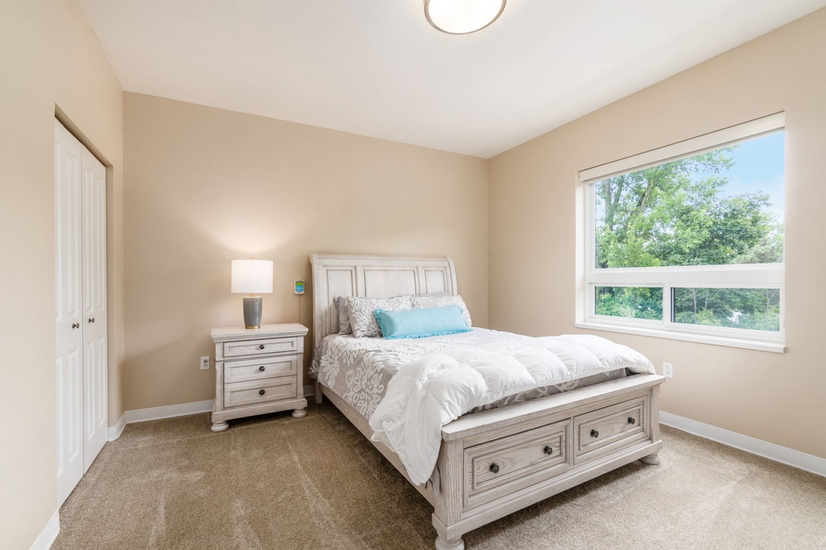 Bright and cozy bedroom unit with a bed, nightstand, lamp, and a large window showing greenery.