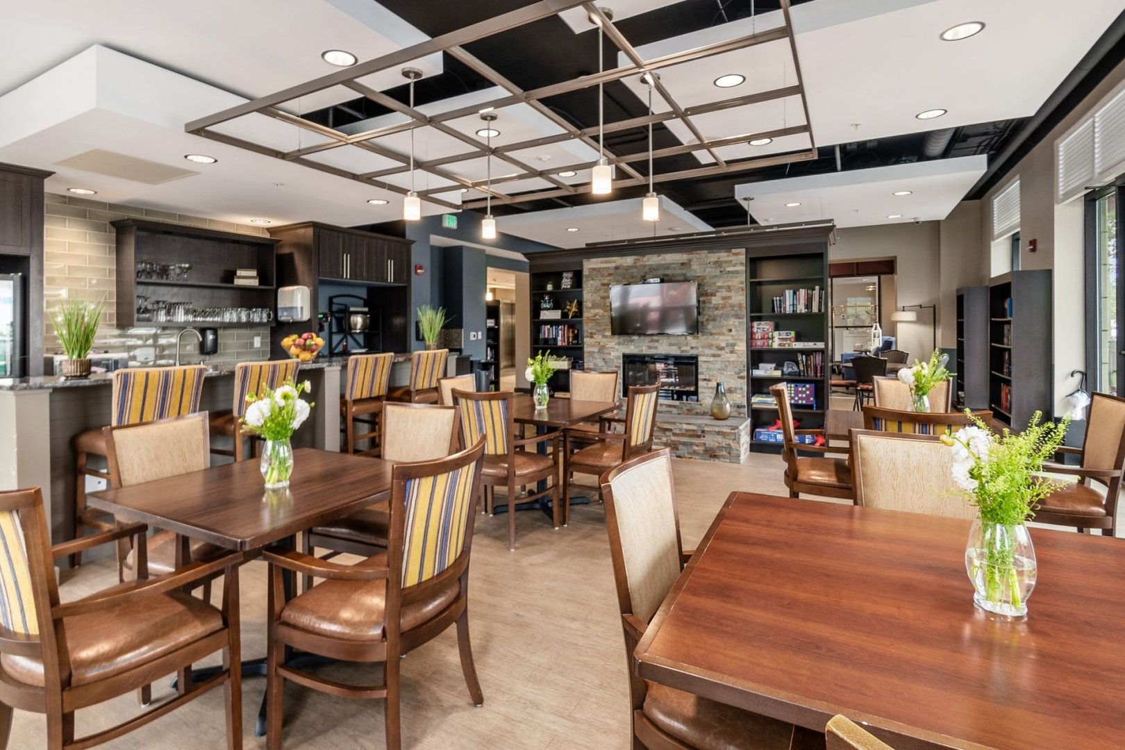 Modern communal dining and lounge area in senior living community with tables, chairs, and a TV.