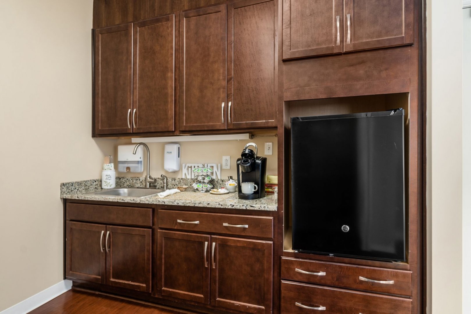 Compact kitchenette with dark wood cabinets, granite countertops, sink, and coffee maker.