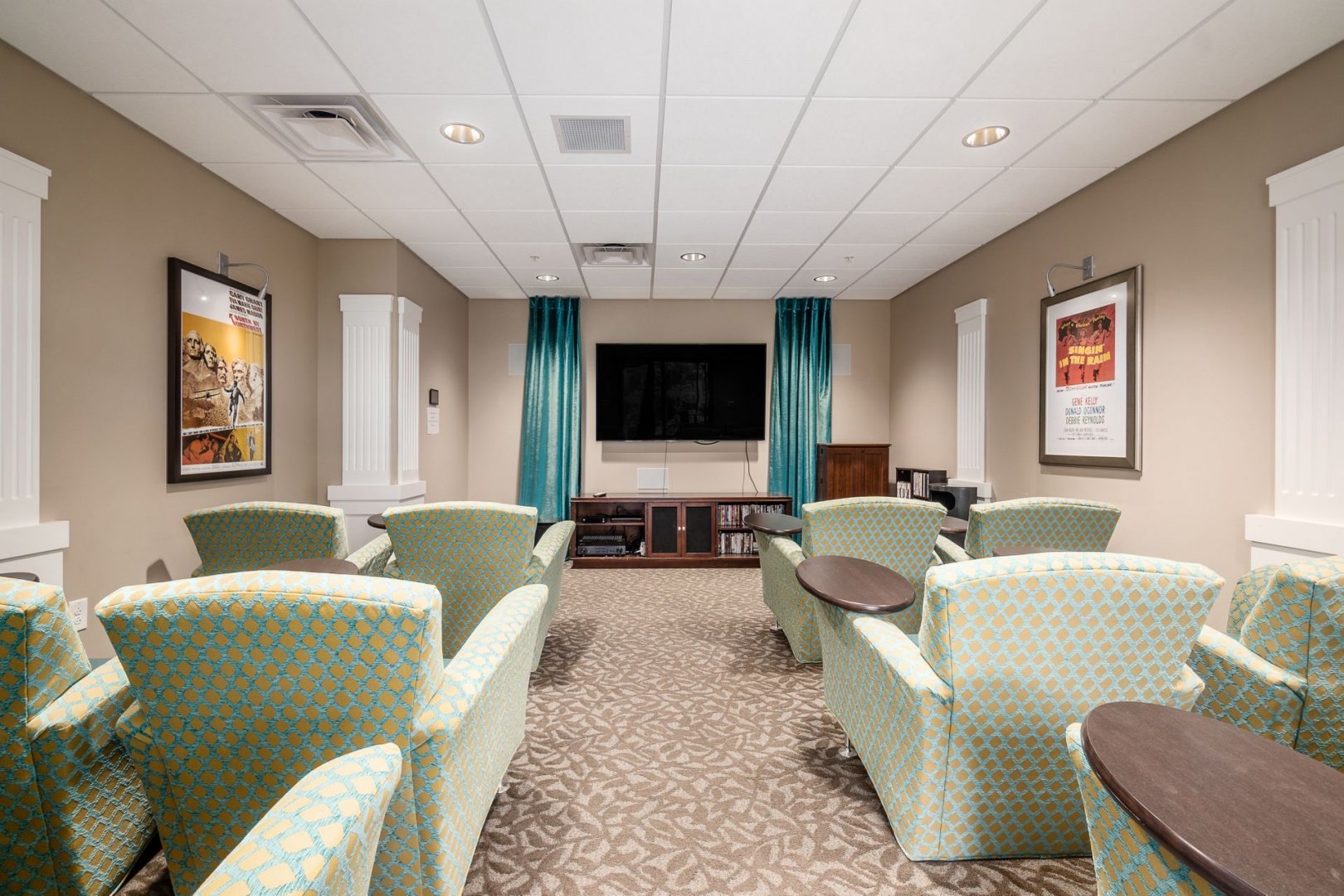 A cozy media room with patterned chairs, a large TV, and posters on the wall in a senior living community.