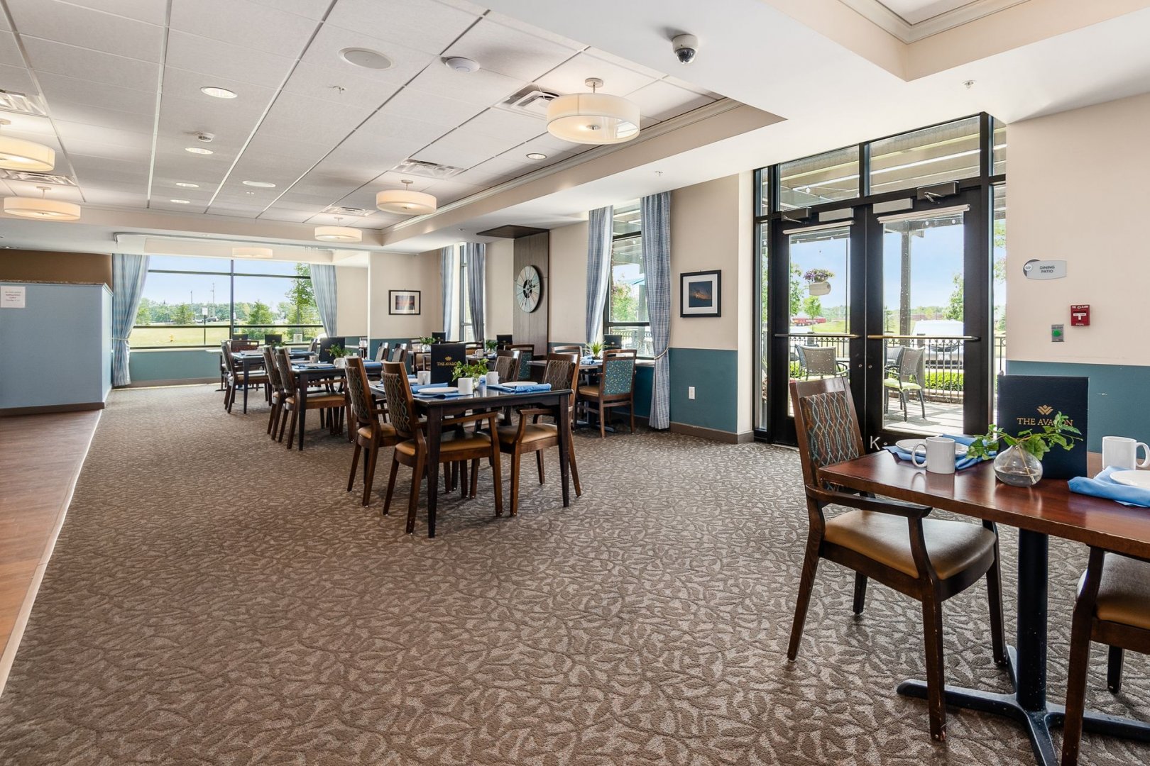Community dining area with multiple tables and chairs, surrounded by large windows and natural light.